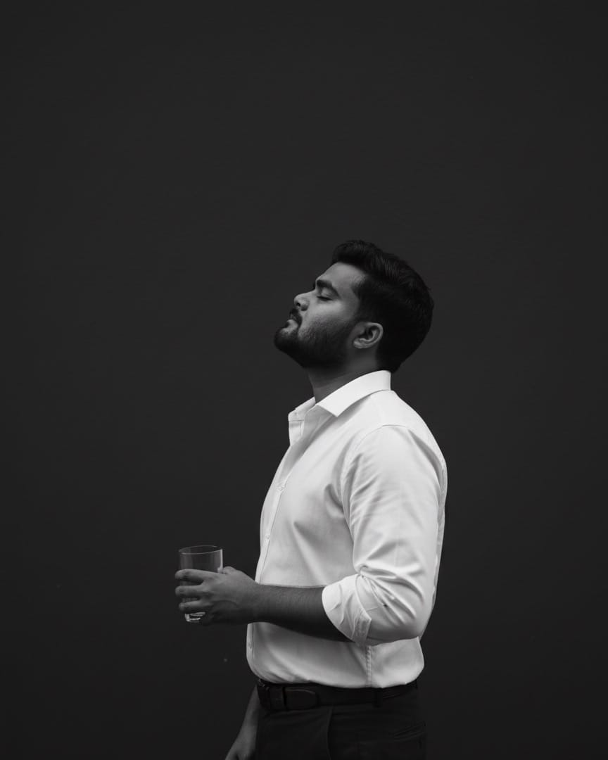 Black-and-white side profile of Aditya against a dark wall, eyes closed, holding a glass.